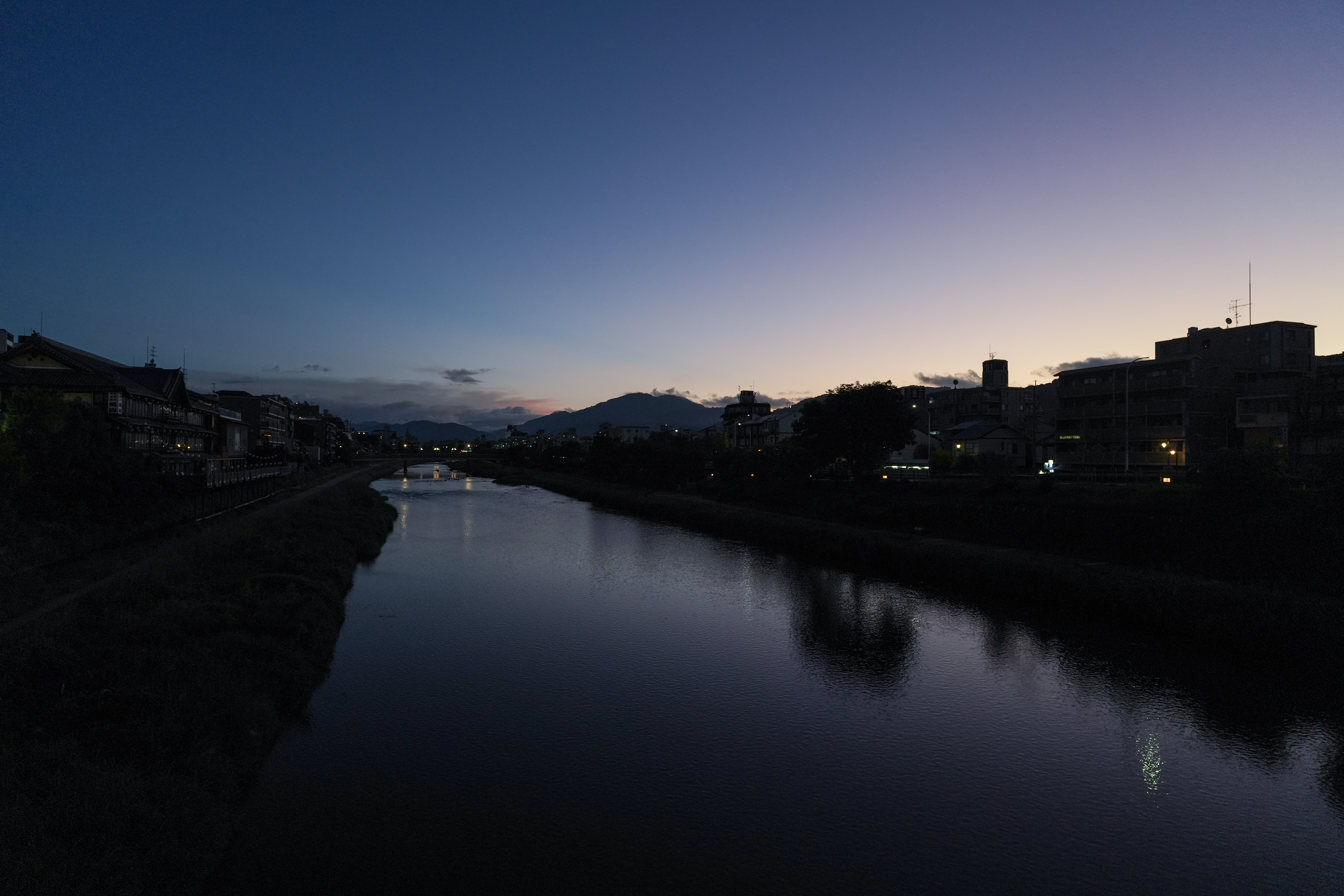 The Kamo River at dusk, with riverside buildings and distant mountains beneath a blue evening sky